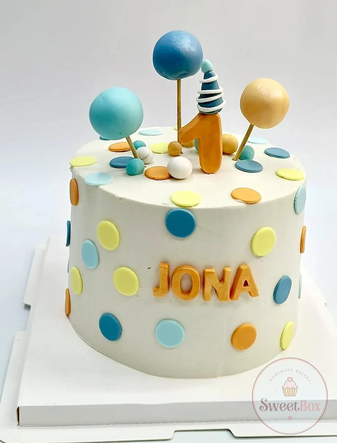 White first birthday cake with pastel polka dots, a party hat number 1 topper and a name in orange letters