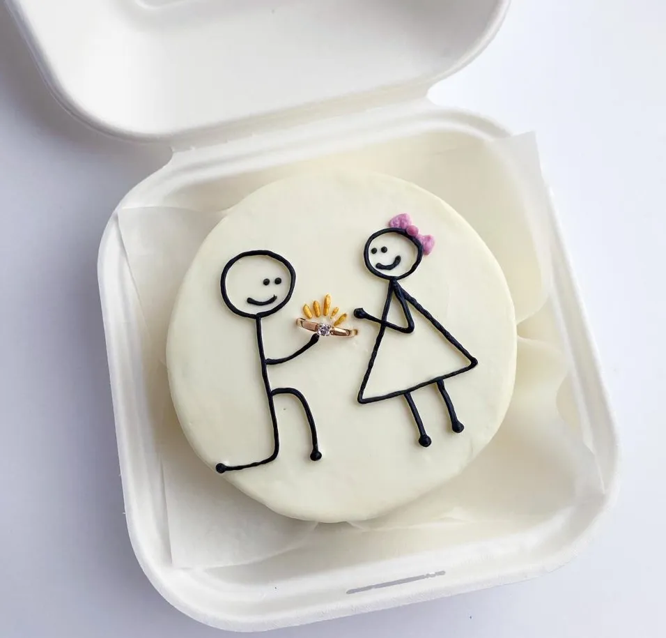 Bento cake in a takeaway box with stick figures of a marriage proposal, a man kneeling with a ring