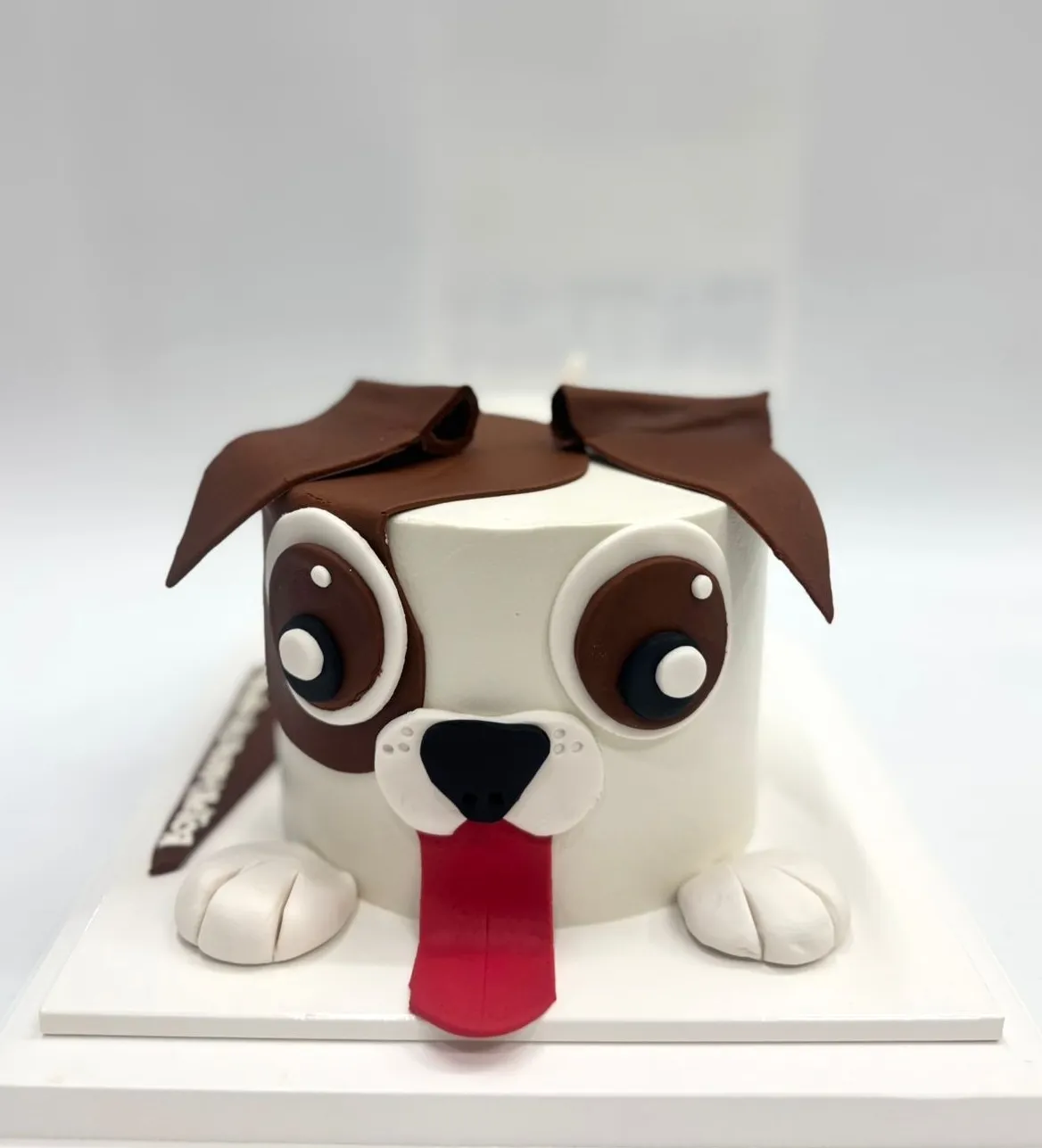 White cake shaped as a puppy face with floppy brown ears, big eyes, white paws and a long red tongue