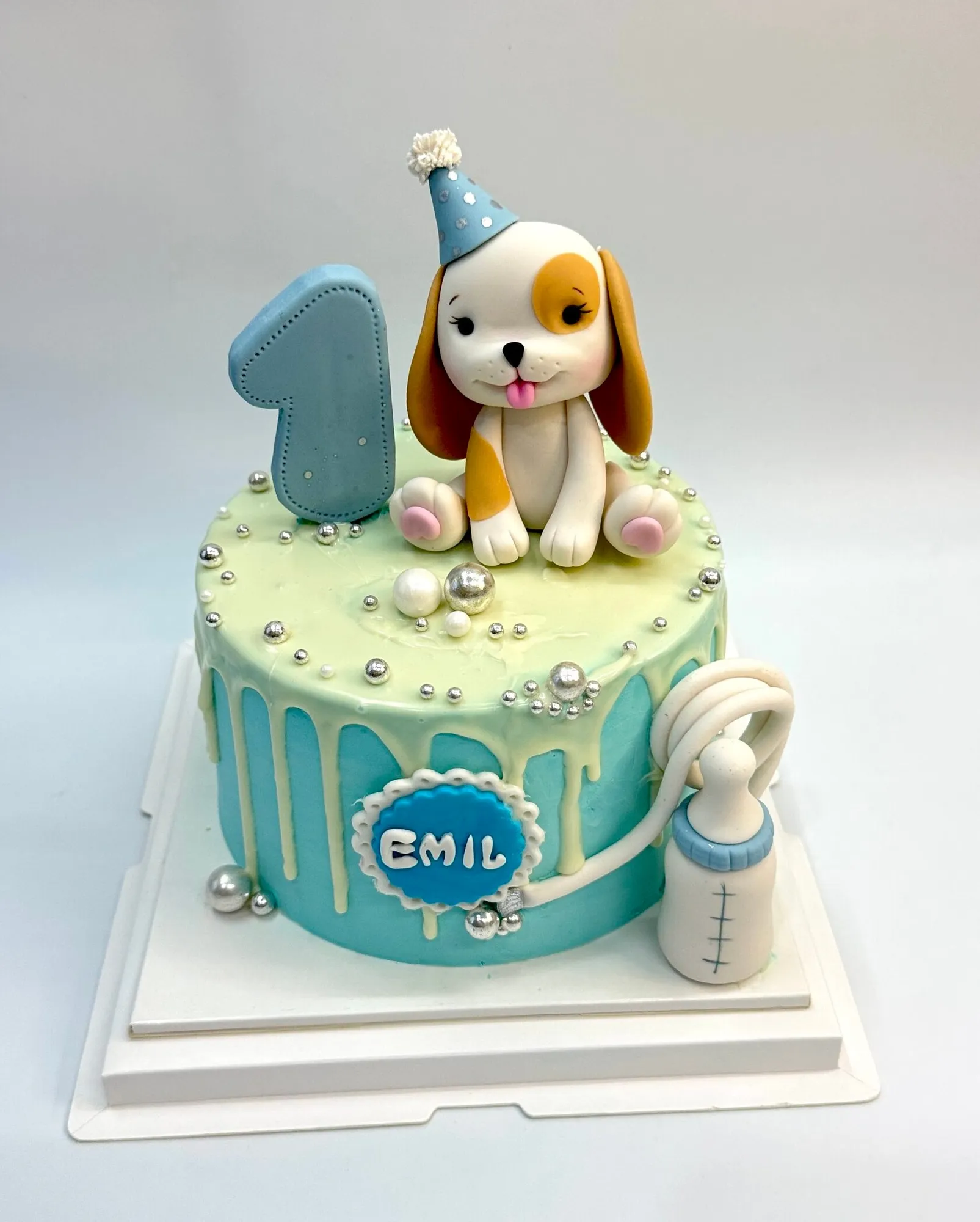 Blue drip first birthday cake with a fondant puppy in a party hat, a number 1, a baby bottle and a name plaque