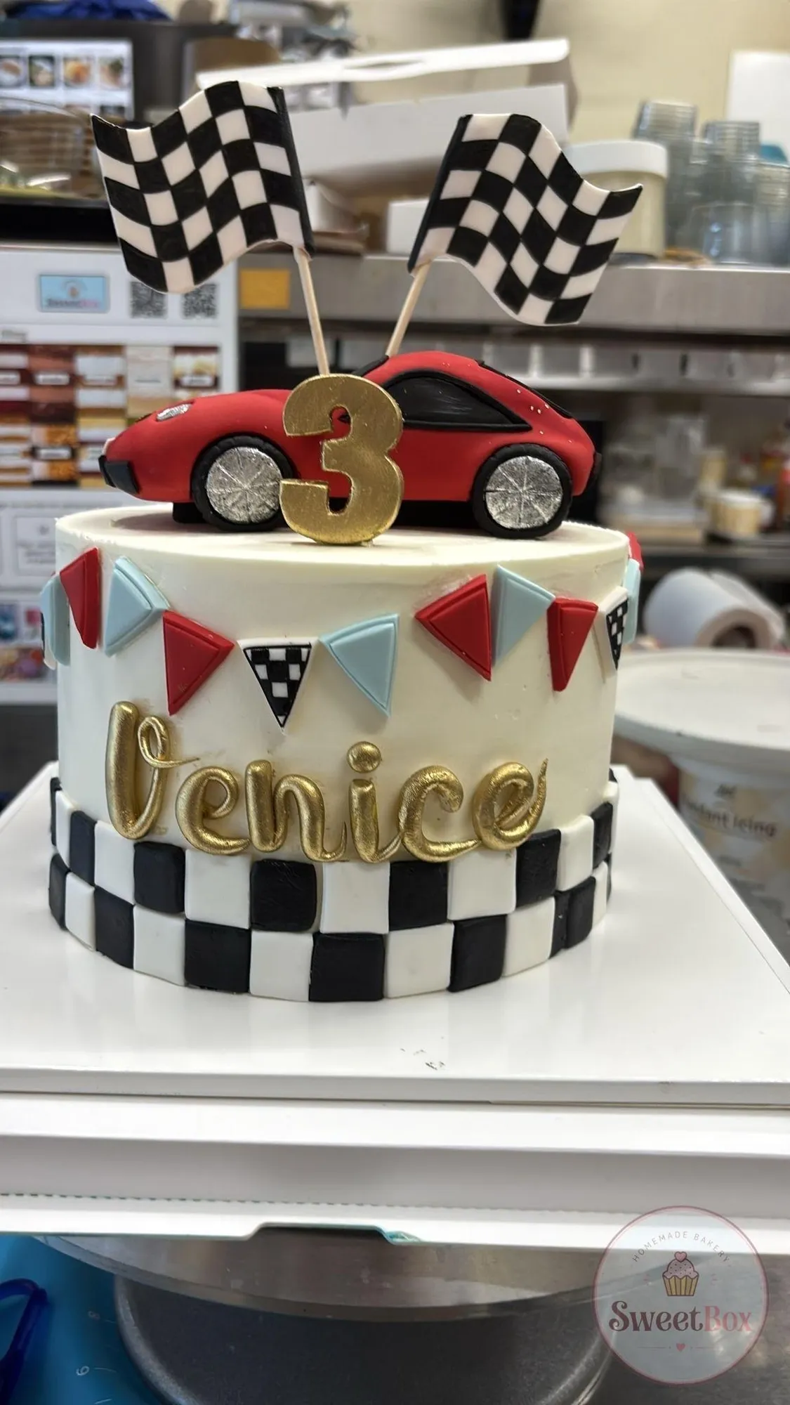 White cake with a red sports car, gold number 3, checkered flags, colorful bunting and a gold Venice name