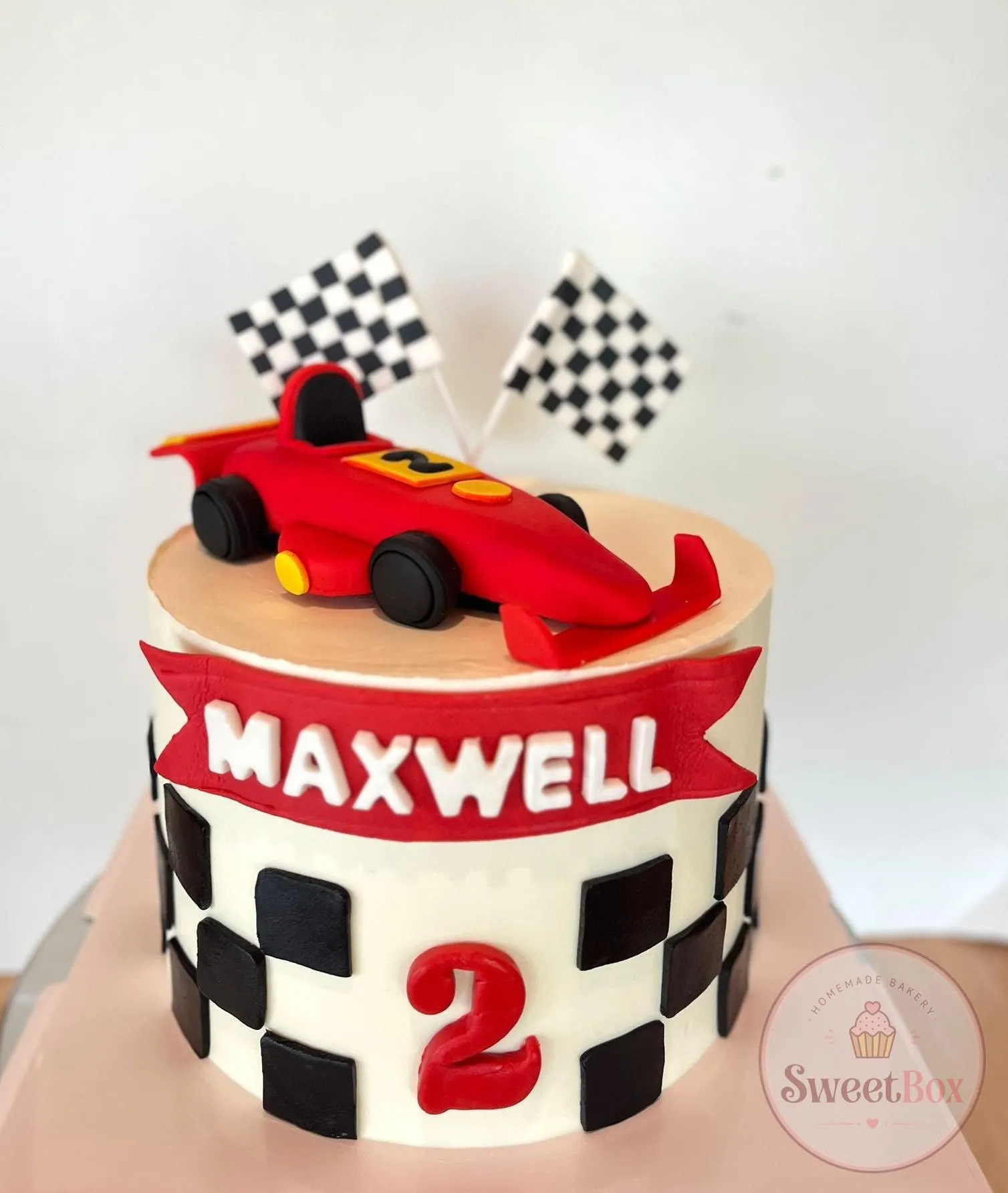 White cake with a red fondant race car, two checkered flags, a red Maxwell banner, black checks and a red number 2