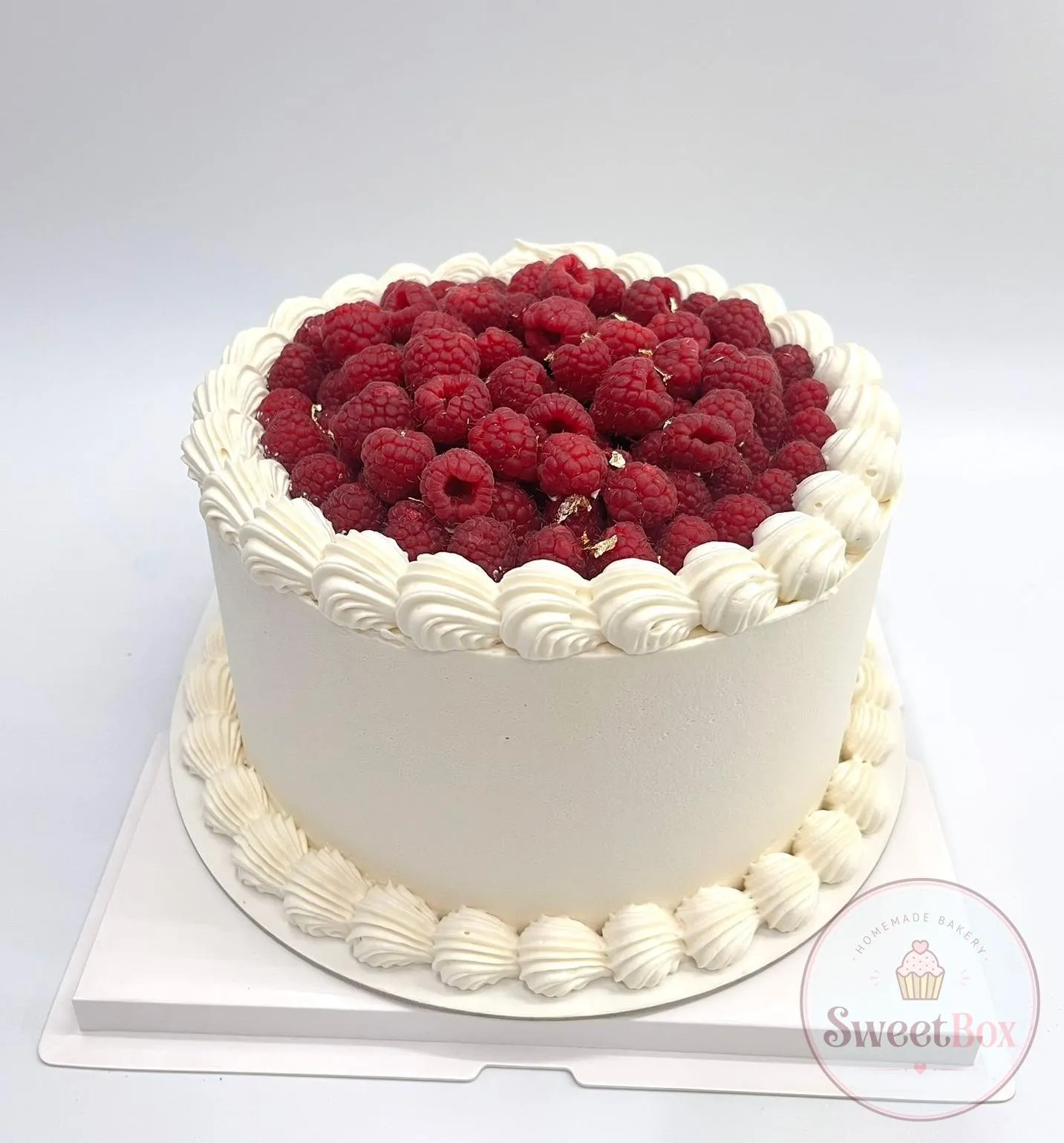 White cream cake with a shell-piped border, its top filled with fresh raspberries and flecks of gold leaf