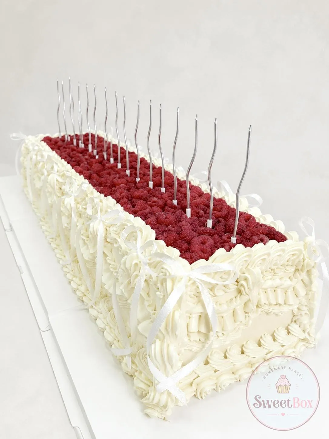 Long rectangular white cake topped with raspberries, cream bows and tall spiral candles