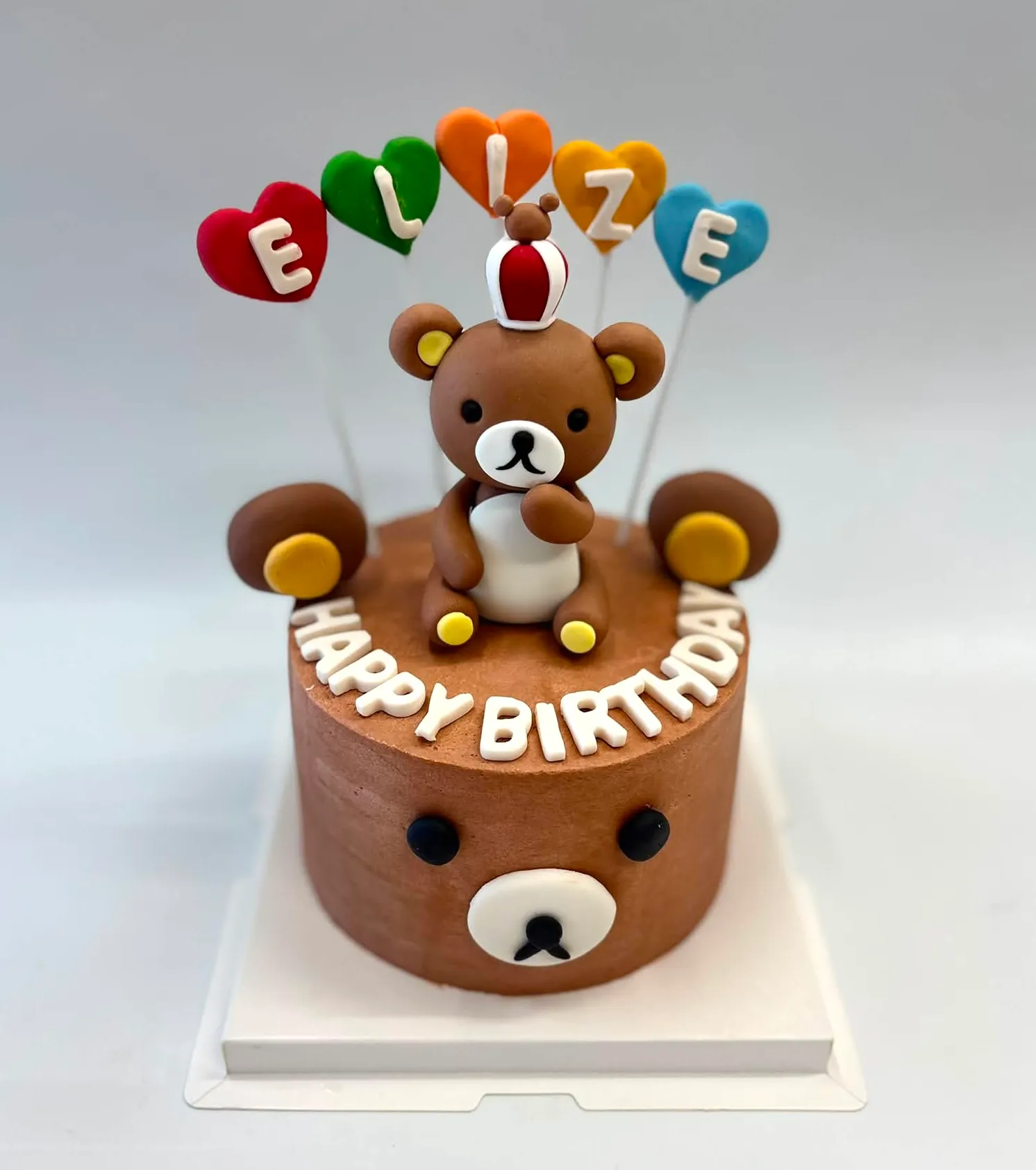 Chocolate bear cake with a fondant bear in a crown, Happy Birthday letters around the edge and a name on colorful heart balloons