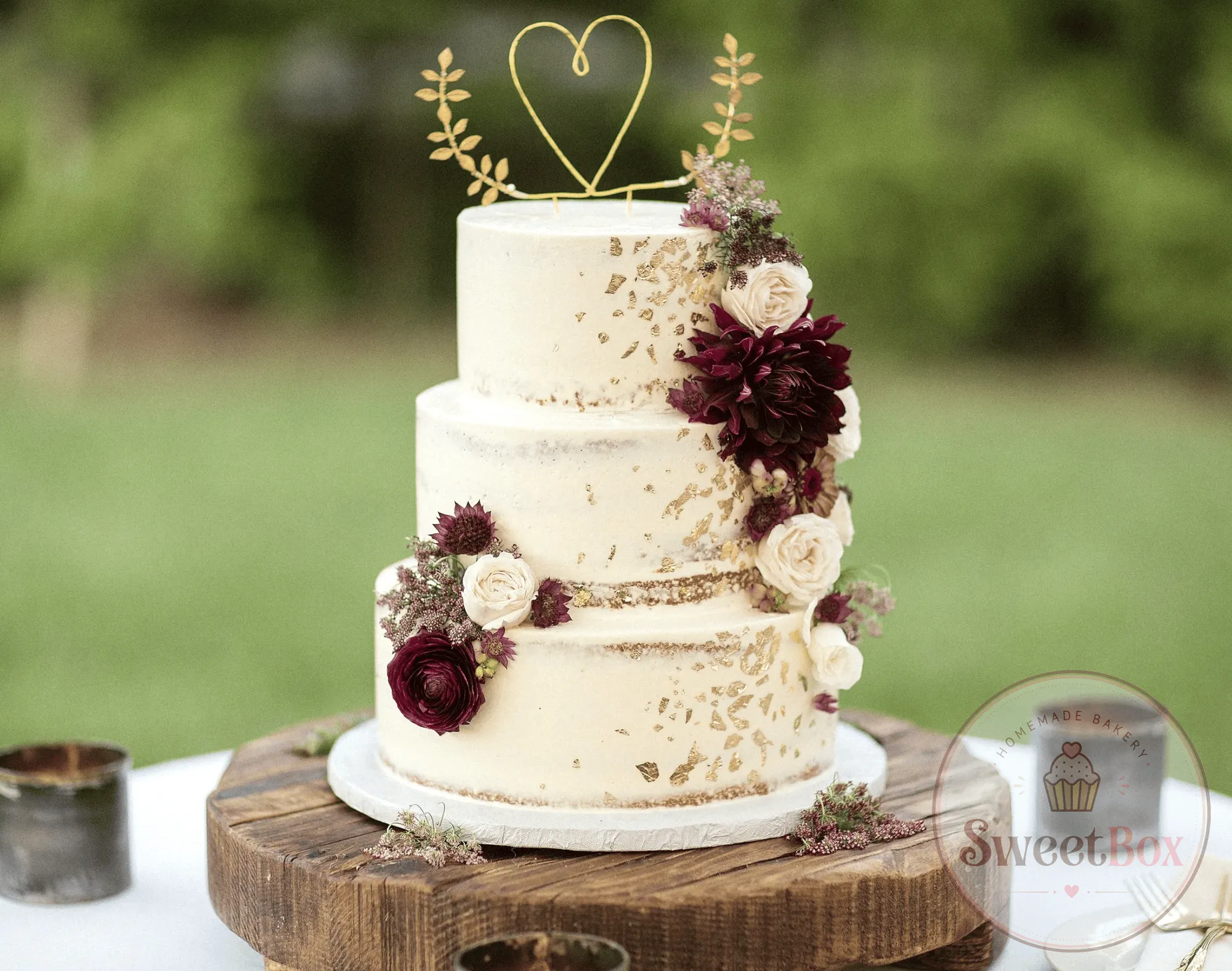 Three-tier semi-naked cake with burgundy and white flowers and a gold heart topper outdoors