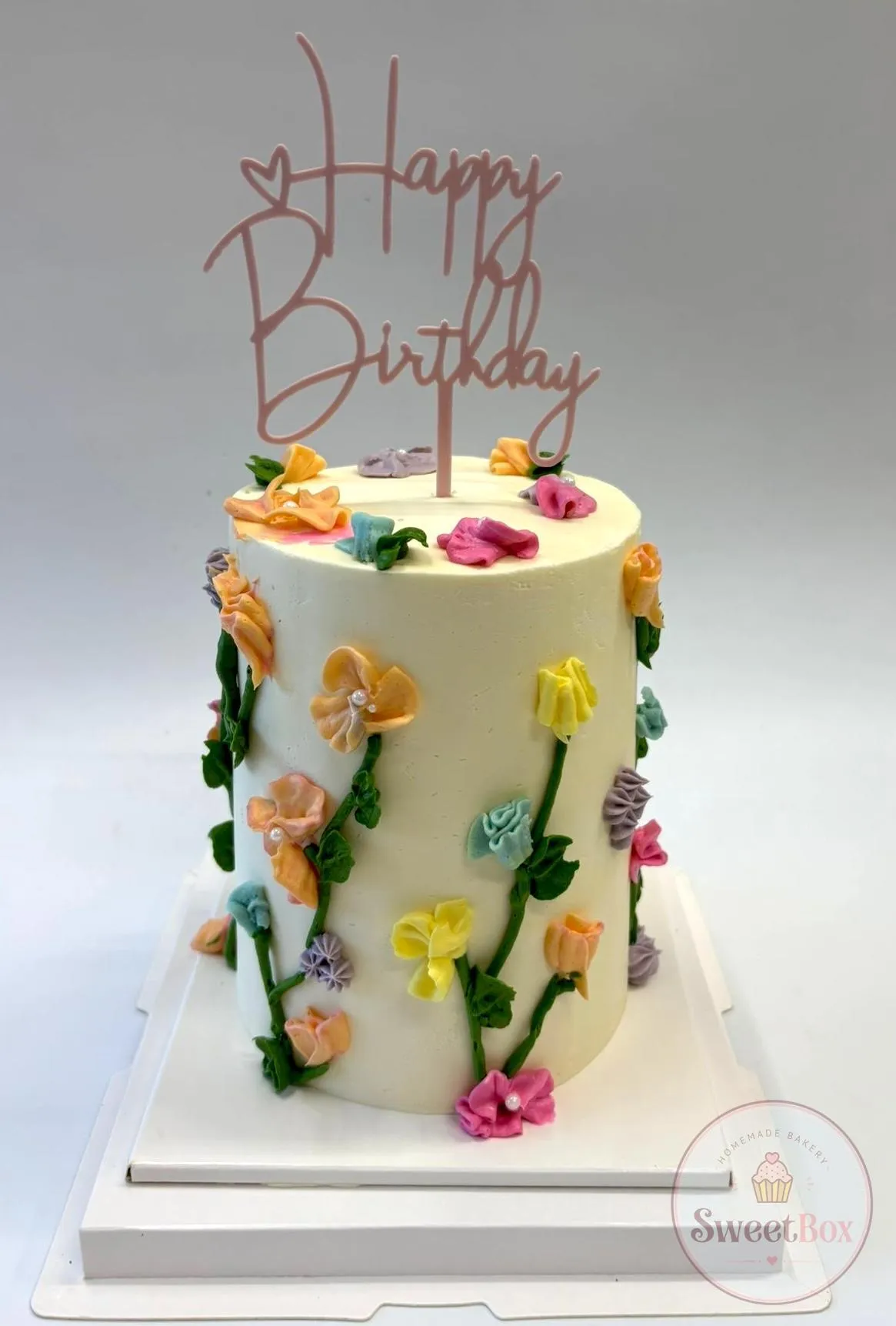 Tall white cake with colorful piped buttercream flowers on green stems and a pink Happy Birthday topper