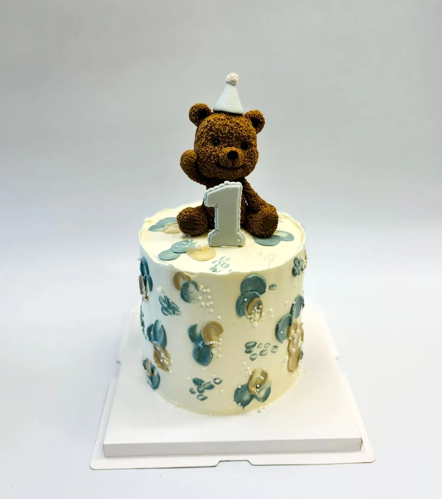 Tall white cake with blue and beige painted dots, a brown teddy bear in a party hat and a number 1 topper