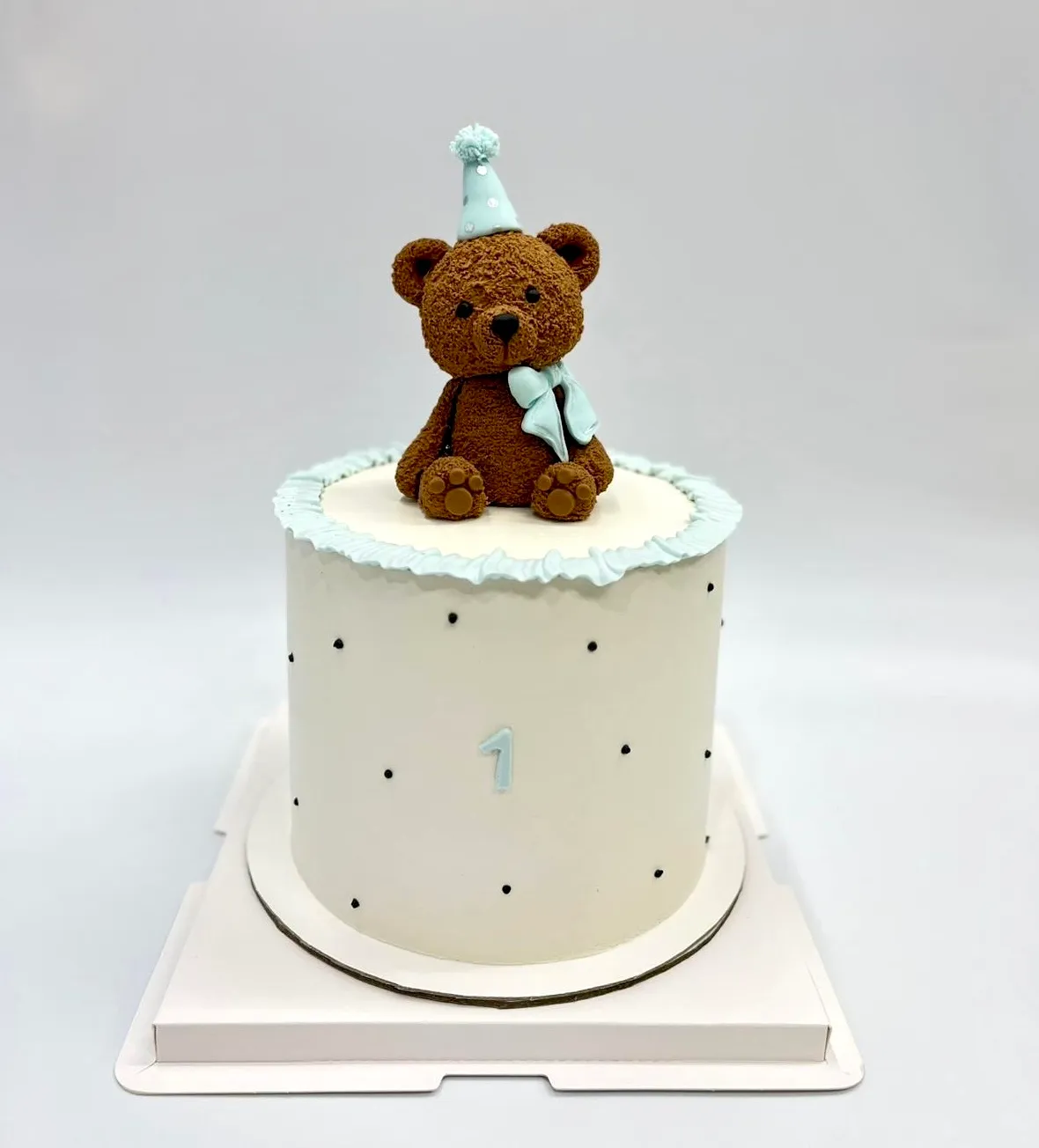 White cake with tiny black dots topped with a brown teddy bear in a blue party hat for a first birthday