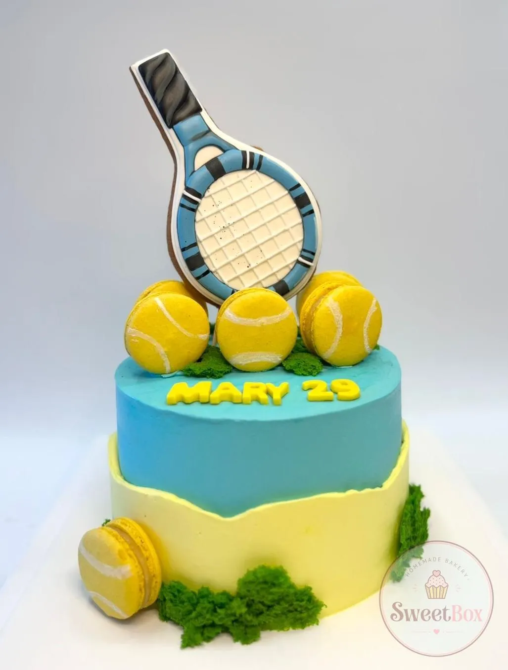 Blue and yellow tennis cake with a decorated racket cookie on top, yellow tennis balls, green crumb grass and a name with the age