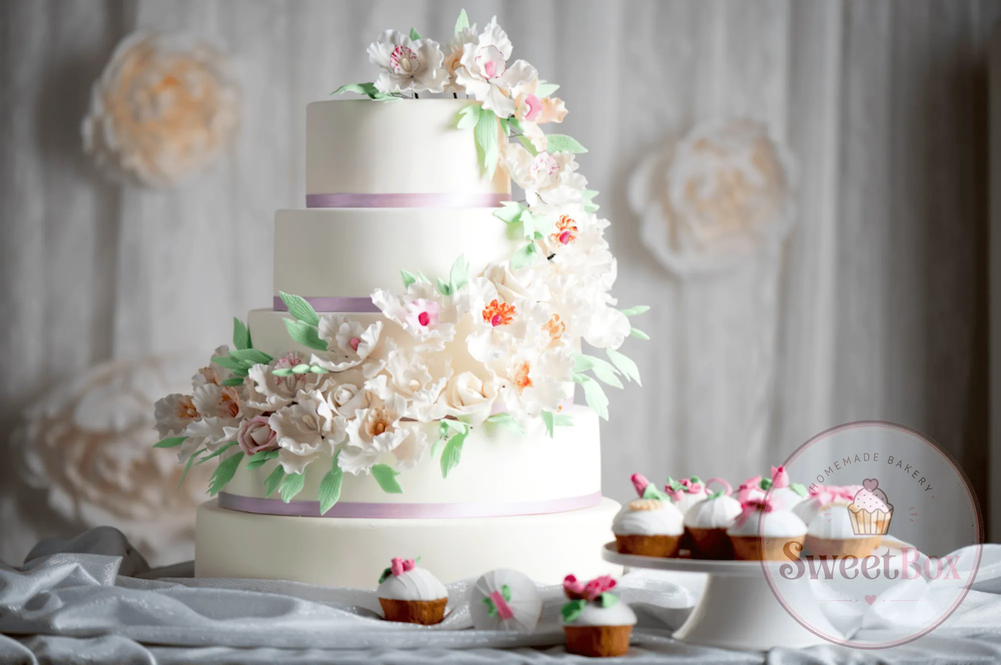 Three-tier white cake with cascading pastel sugar flowers and lilac ribbons beside cupcakes