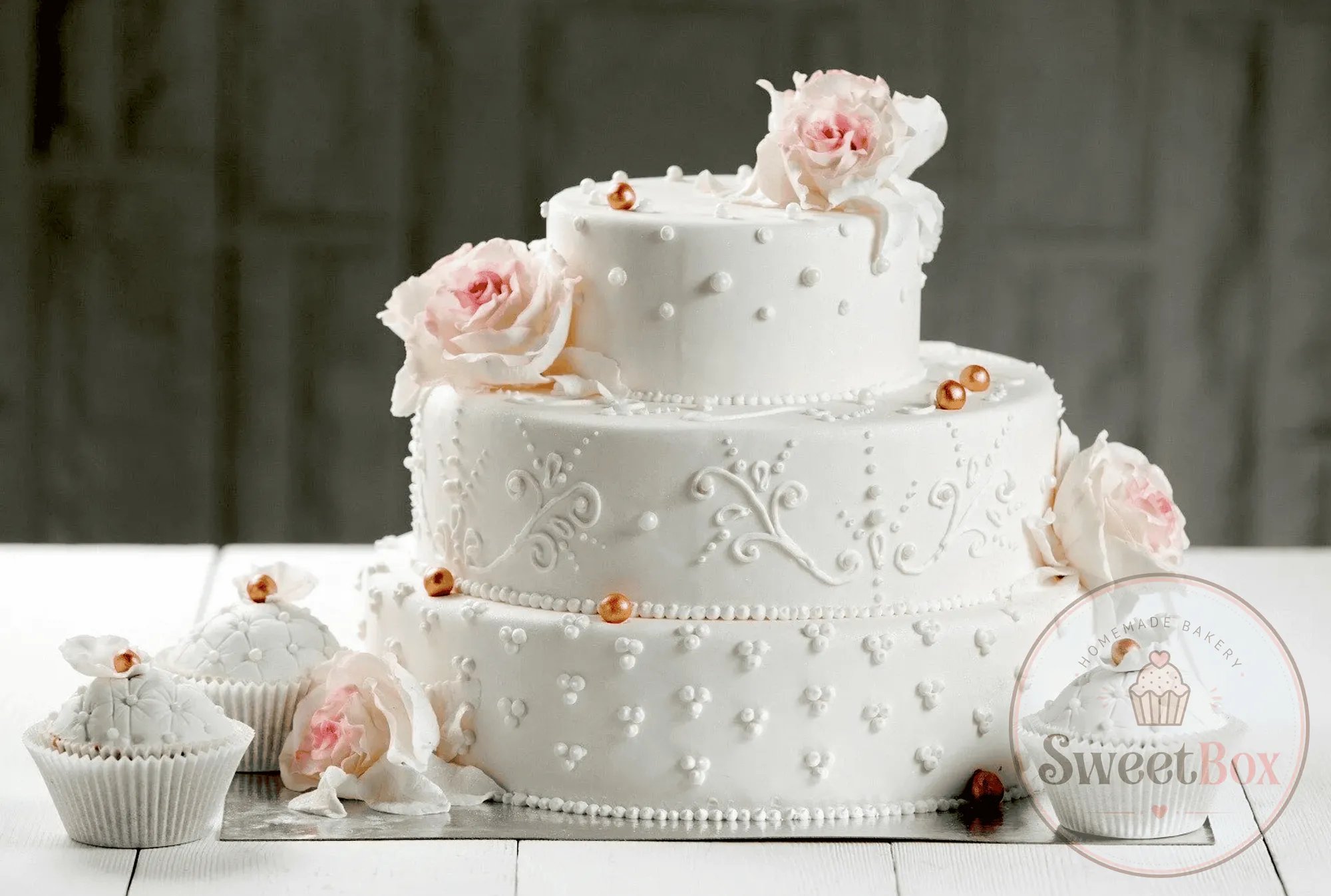 Three-tier white cake with lace piping, pearls and pink roses next to matching cupcakes
