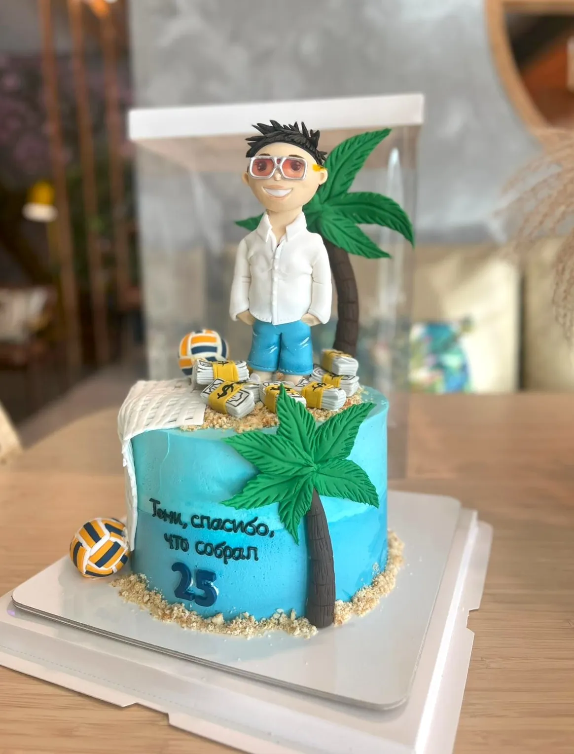 Turquoise beach-themed birthday cake with a fondant man in sunglasses, palm trees and a number 25 in a clear box