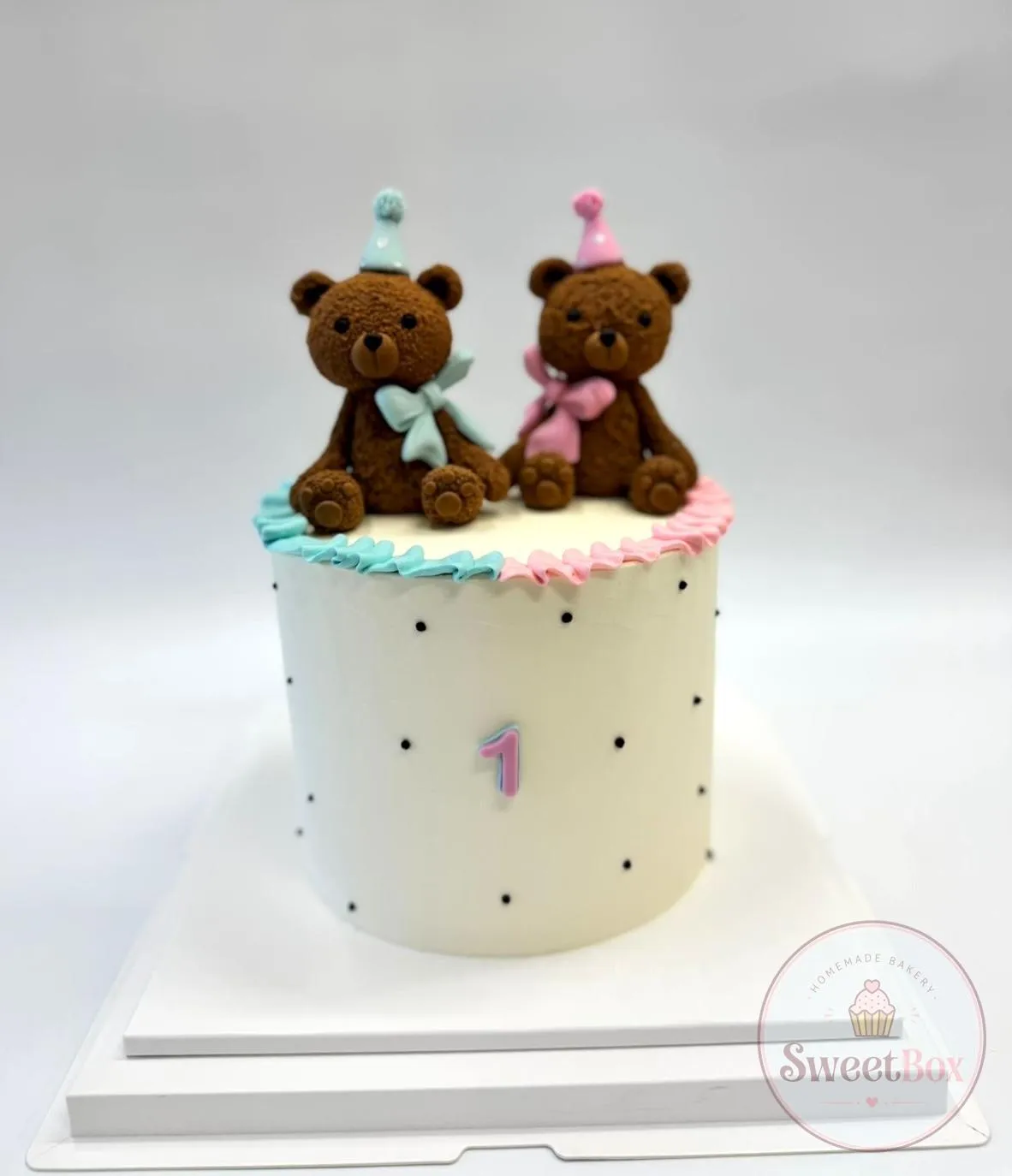 White first birthday cake with black dots, two teddy bears in blue and pink party hats and a small number 1