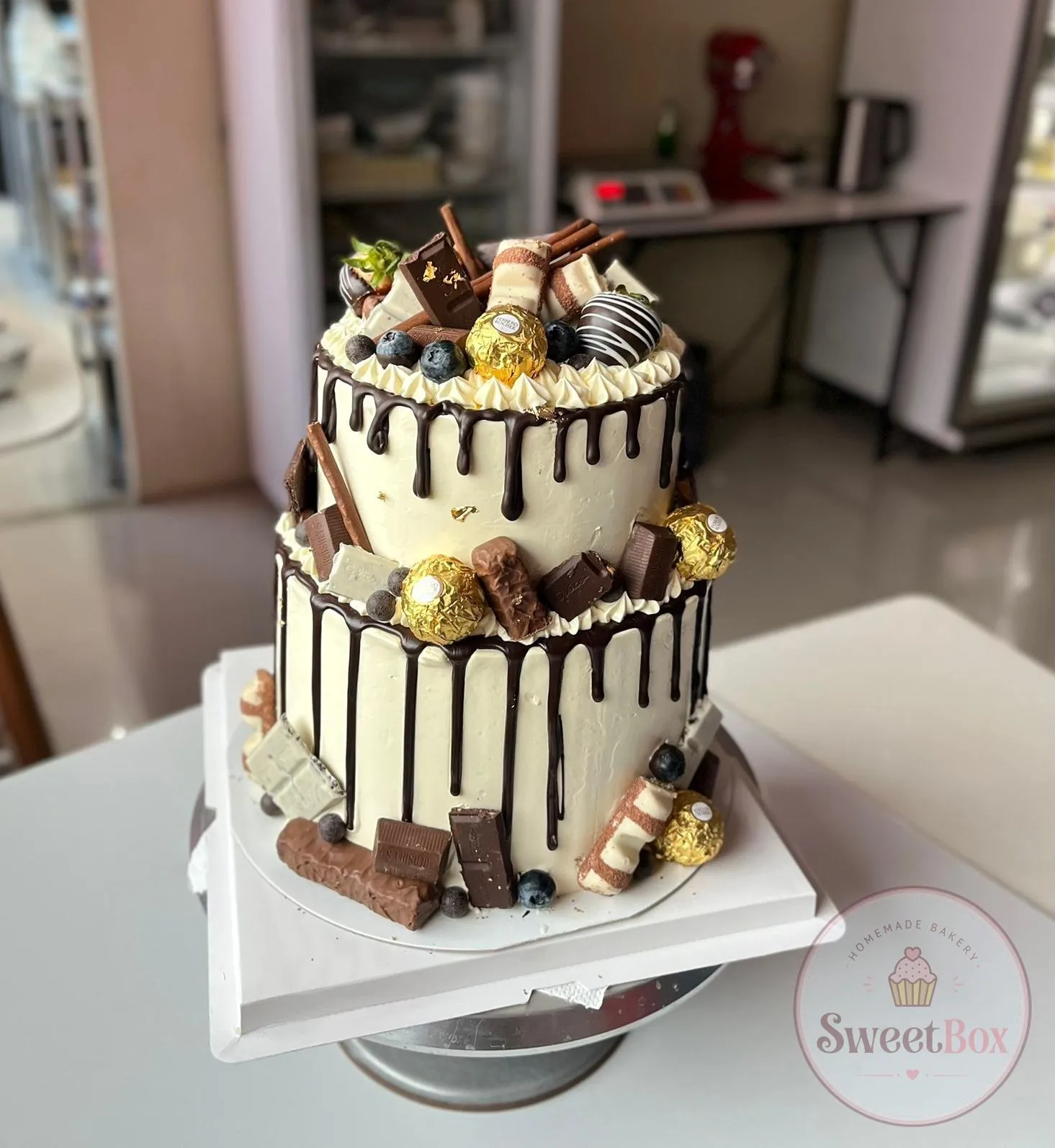 Two-tier white cake with dark chocolate drip, chocolate bars, gold-wrapped truffles and blueberries