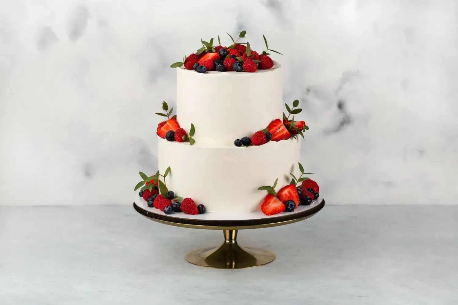 Two-tier white wedding cake decorated with fresh strawberries and blueberries on a gold stand