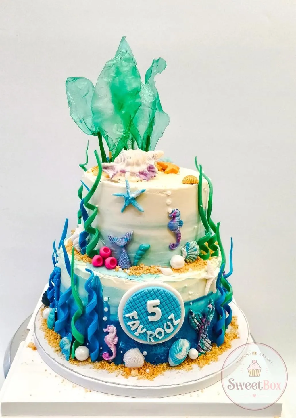 Two-tier ocean cake with sugar seaweed sails, coral, seahorses, shells, pearls and a blue plaque with number 5 and a name