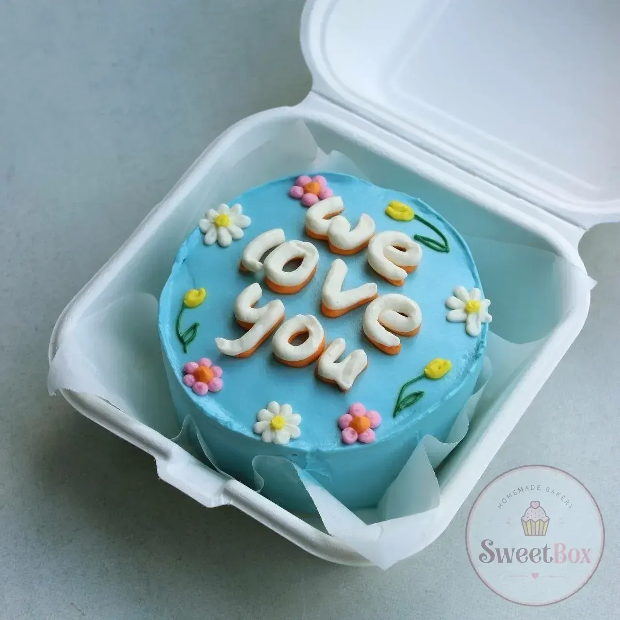 Sky blue bento cake with We Love You lettering and small piped daisies and tulips in a takeaway box