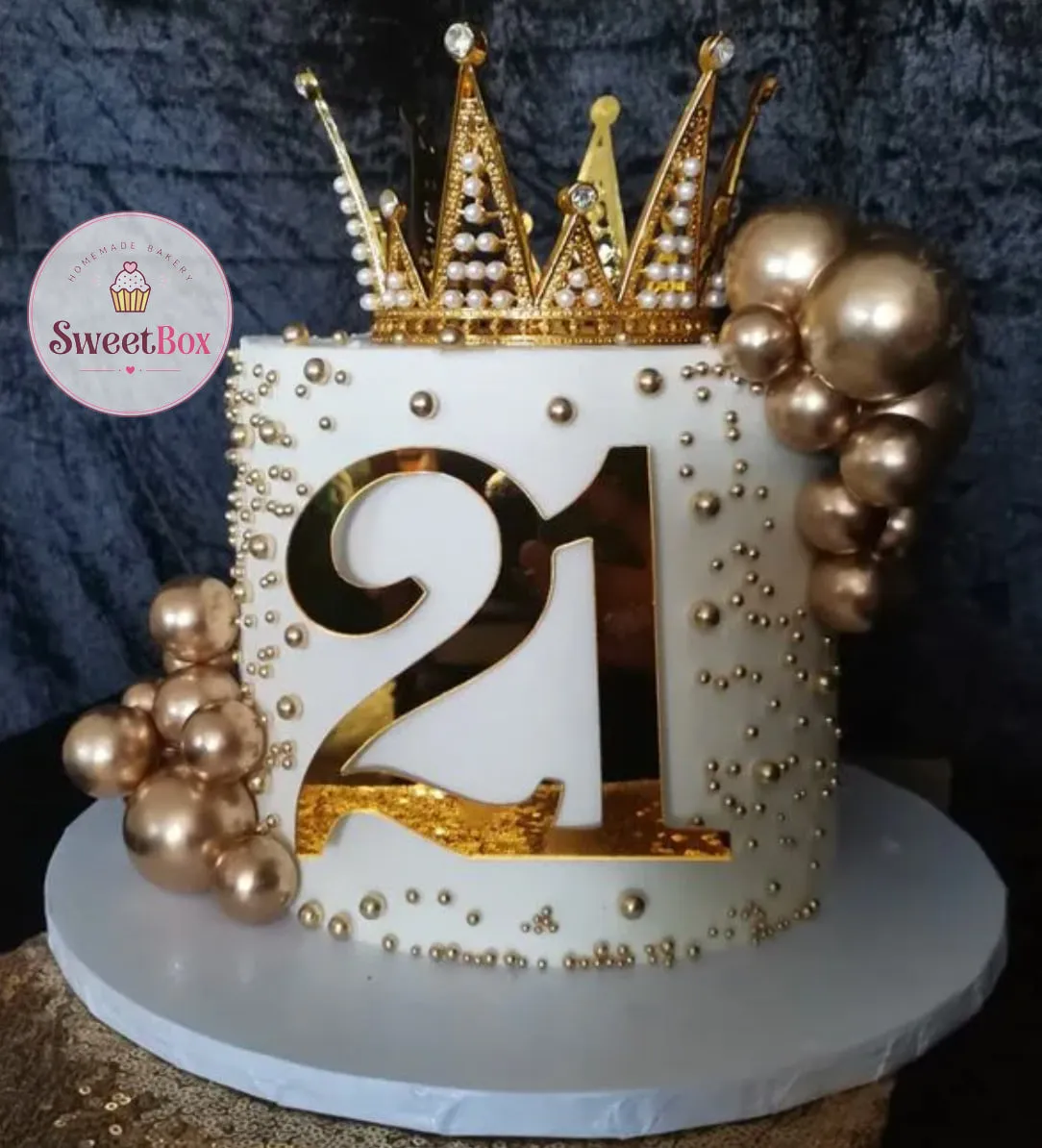 White cake with a large mirror-gold number 21 on the side, a gold crown with pearls on top, gold spheres and scattered gold pearls