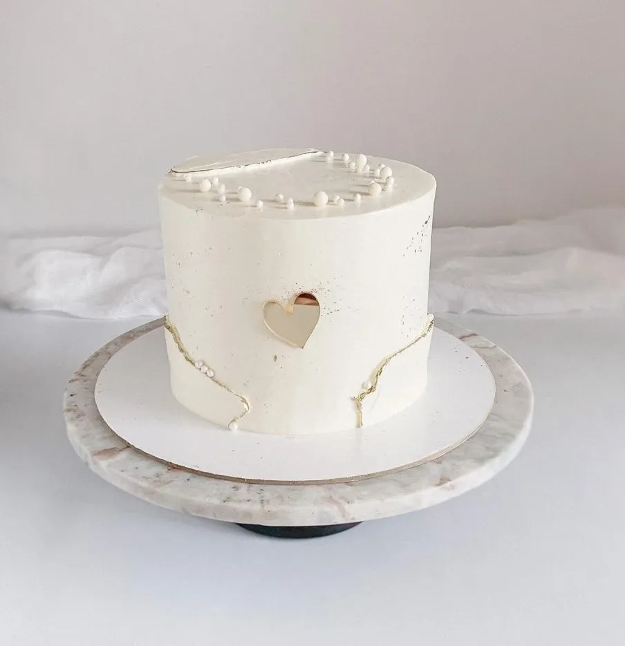 Minimal white cake with a heart-shaped cutout, gold crack details and white pearls on a marble stand
