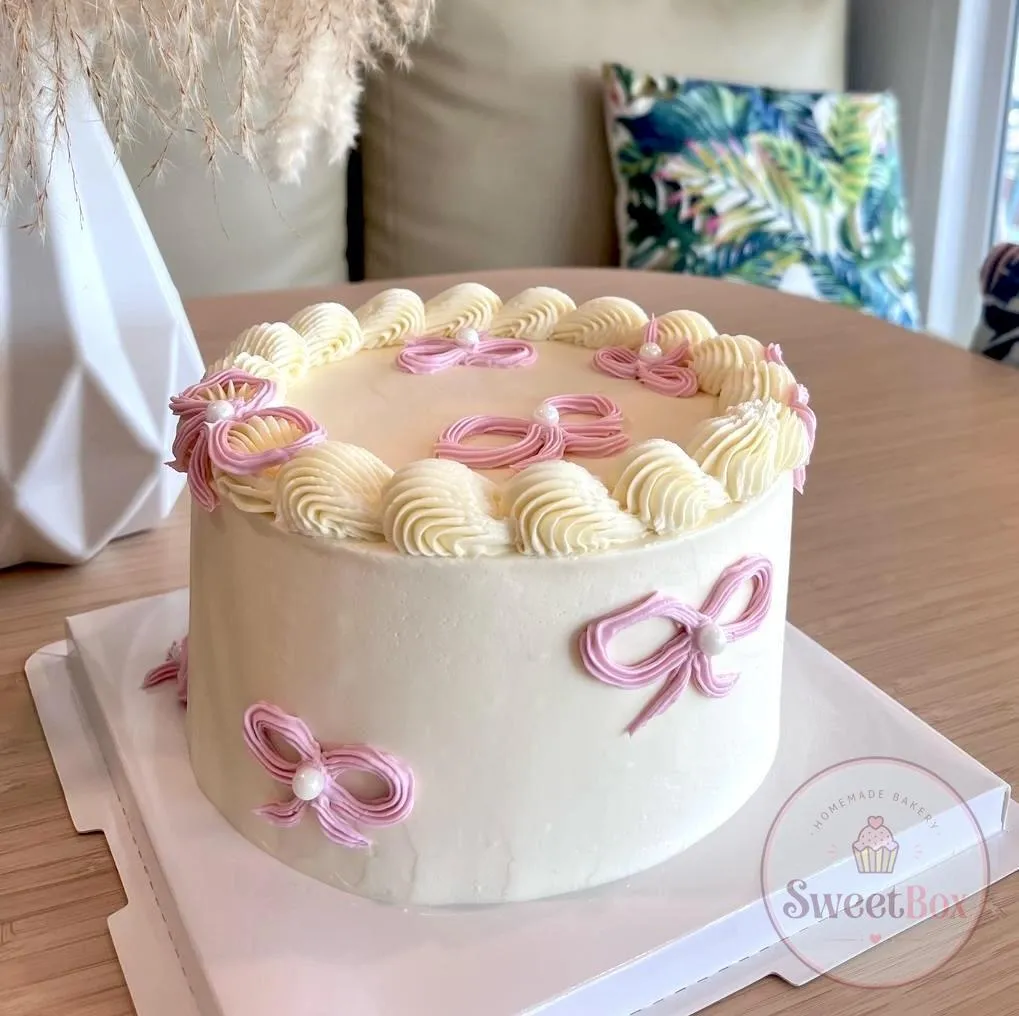 White cake with a cream rosette border and pink piped bows, set on a table beside pampas grass