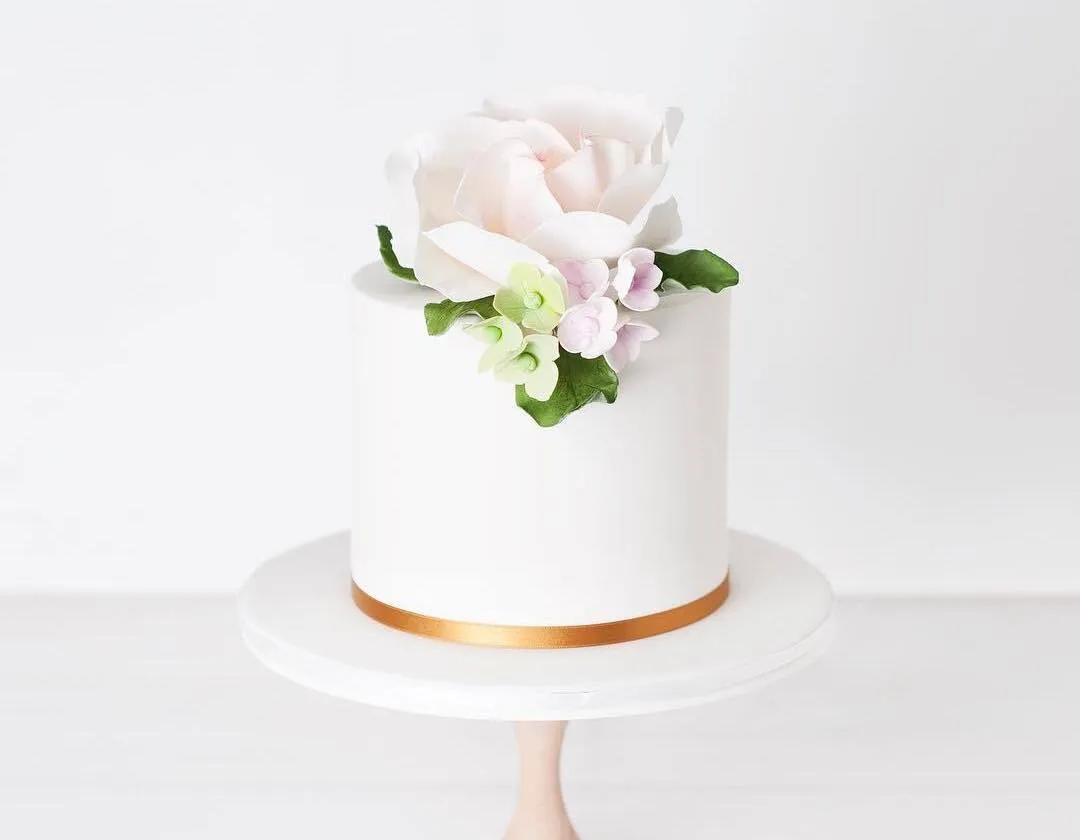 Single-tier white cake with a sugar peony, green leaves and a gold ribbon base